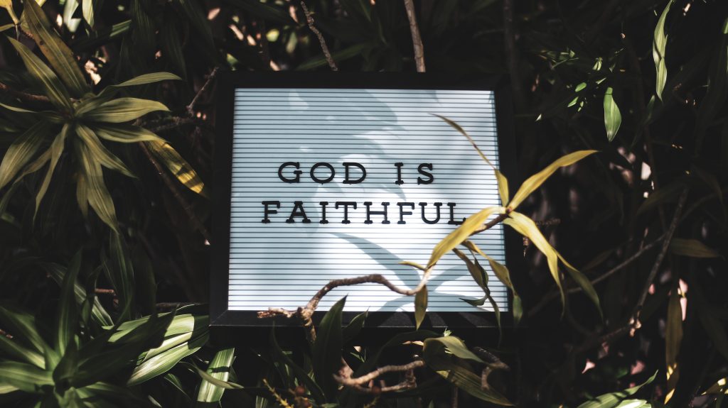 A letter board reading 'God is Faithful' set among leafy plants.