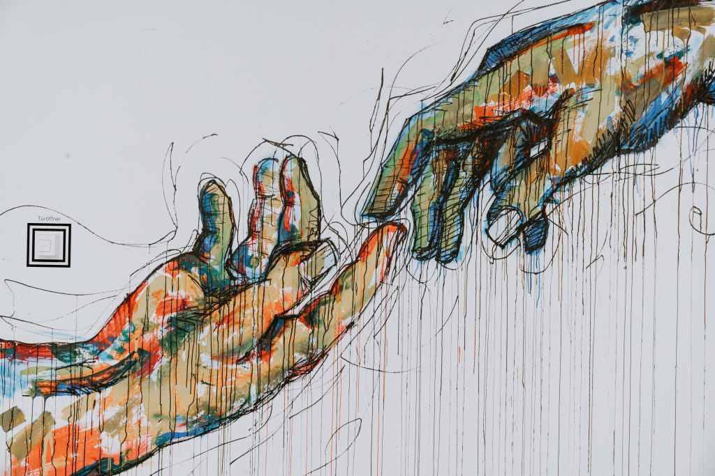A painted sketch of two hands reaching toward each other, echoing Michelangelo's Creation of Adam, with drips of colored paint.