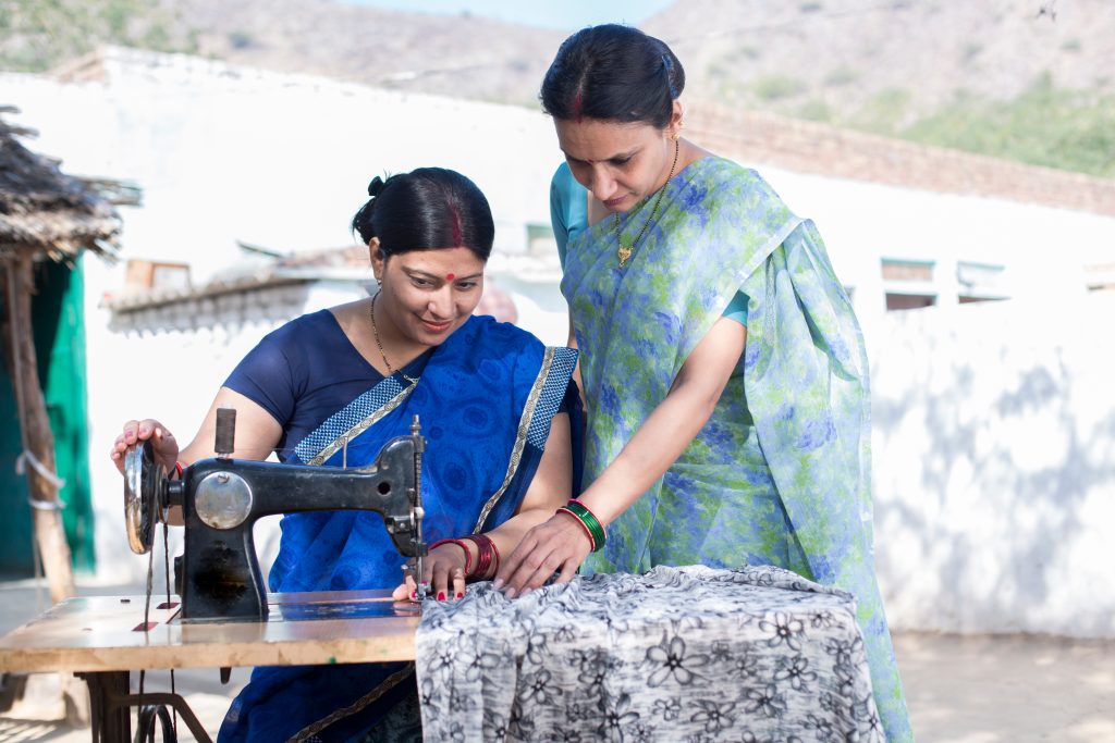 Two women in saris work together at a manual sewing machine outdoors.